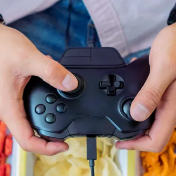 Person hält einen Gamecontroller über Schalen mit Snacks (Chips und Knabberzeug).
