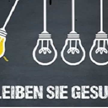 Eine leuchtende Glühbirne stößt andere nicht glühende an. Schriftzug darunter: Bleiben Sie gesund