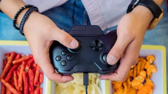 Person hält einen Gamecontroller über Schalen mit Snacks (Chips und Knabberzeug).